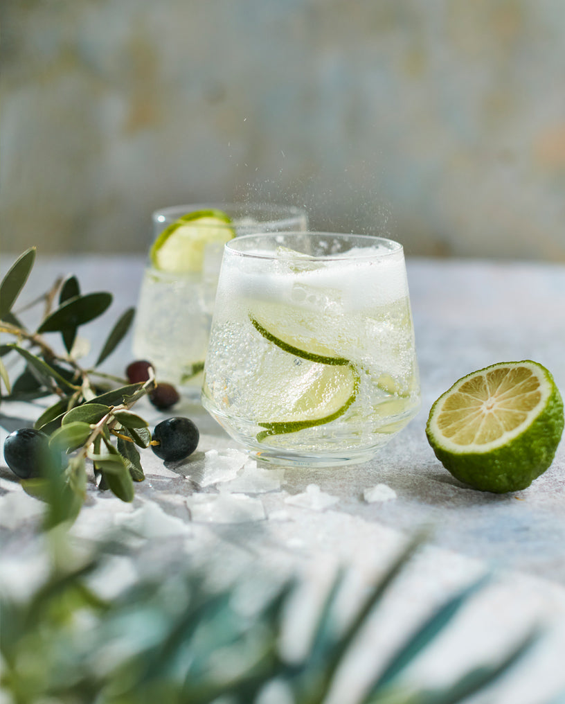 Two glasses filled with Everleaf’s Marine and Tonic, surrounded by a slice of lime and olive leaf.